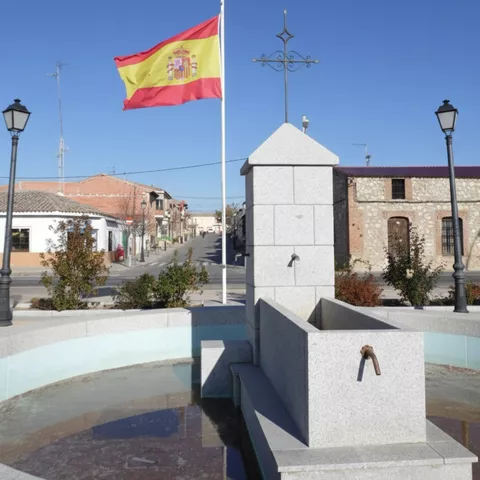 Primer plano detallado de una fuente pública de piedra de granito con grifos de metal y un pilón de agua, flanqueada por farolas y una bandera nacional, con una vista de una calle de pueblo al fondo.