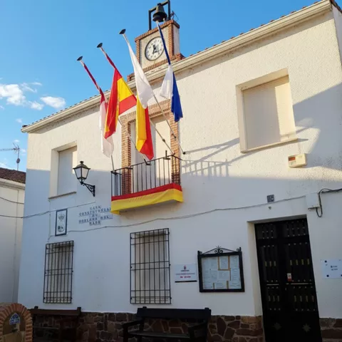 Fachada de un edificio municipal de color blanco con un balcón central decorado con banderas y una bandera colgante. E