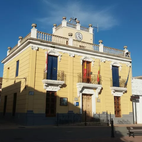 Fachada principal de un edificio municipal de color amarillo con balcones de forja, banderas y un reloj en la parte superior.