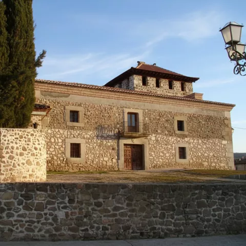Un edificio histórico de piedra con una torre cuadrada, rodeado por un muro bajo y cipreses altos bajo un cielo despejado.