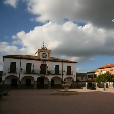 Plaza de pueblo con una fuente central, un edificio blanco con soportales a la izquierda