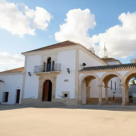 Edificio blanco con arquería lateral y cielo con nubes sobre una explanada.