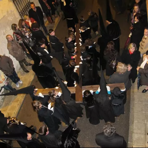 Procesión nocturna con personas vestidas de negro portando una cruz por una calle estrecha.