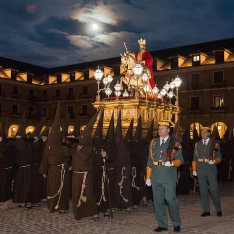 Paso nocturno con imagen religiosa a caballo escoltado por nazarenos y músicos en plaza iluminada.