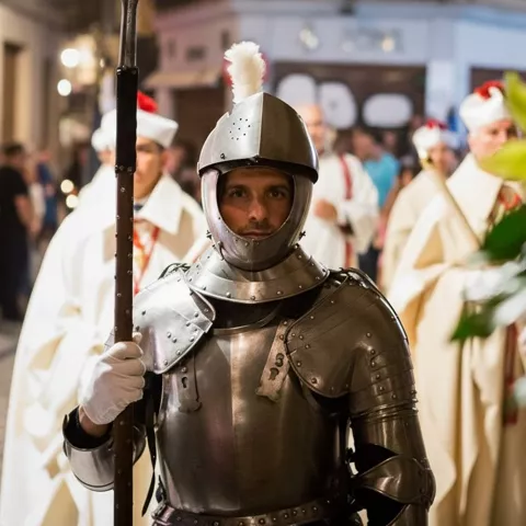 Hombre con armadura medieval y lanza encabezando una procesión nocturna con túnicas claras.