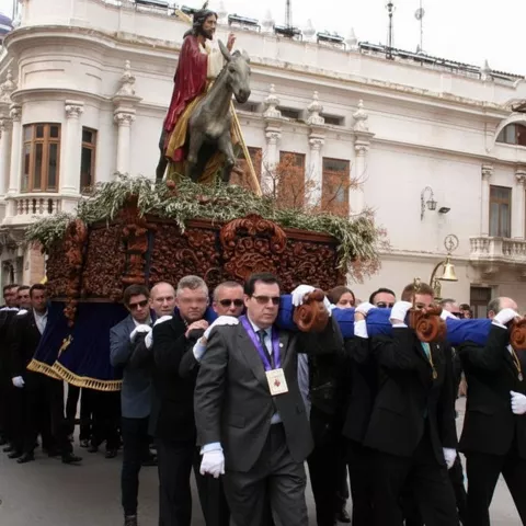 Grupo de costaleros portando un paso con figura religiosa montada sobre un burro, en una plaza con espectadores.