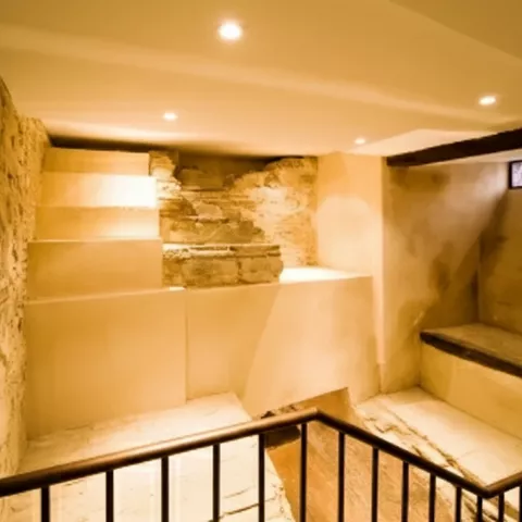 Escalera interior con muros de piedra y restos arqueológicos iluminados en sótano histórico.