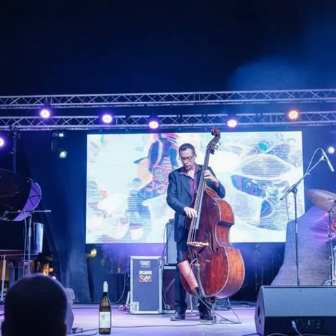 Trío de jazz en escenario con piano, contrabajo y batería bajo luces moradas.