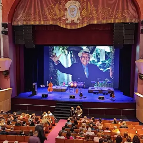 Escenario de teatro con gran pantalla y público ocupando la platea.