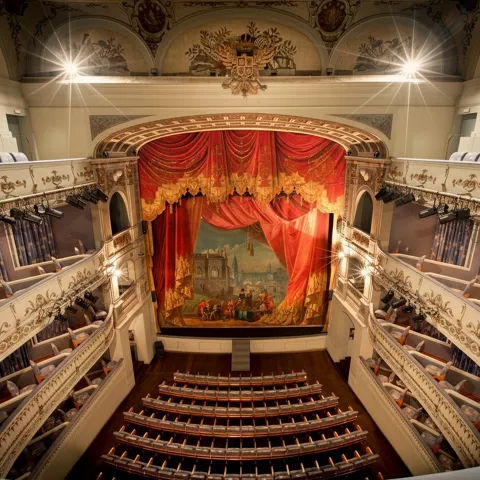 Escenario con telón rojo y balcones laterales en auditorio clásico.