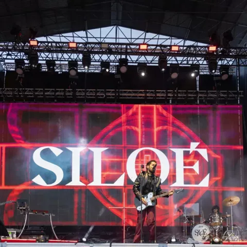 Banda de música actuando en un gran escenario con pantallas y luces rojas.