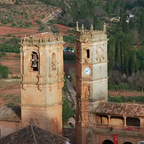 Torres de piedra con campana y reloj sobresaliendo entre tejados, con paisaje rural al fondo.