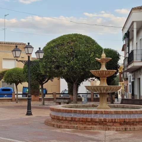 Una plaza adoquinada con una fuente de piedra de tres niveles en primer plano.