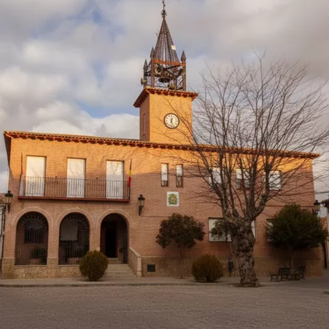 Un ayuntamiento de ladrillo con un campanario y un reloj, con banderas colgando de la fachada y un gran árbol desnudo en primer plano.