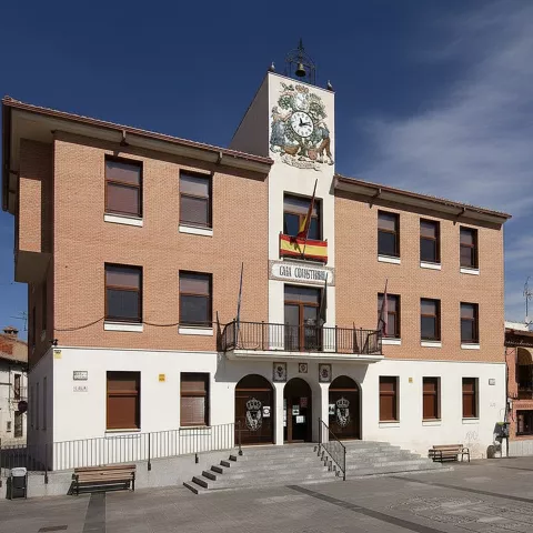 Edificio municipal de tres plantas con ladrillo visto, una hilera de banderas en el balcón central y un gran reloj decorativo en la parte superior.