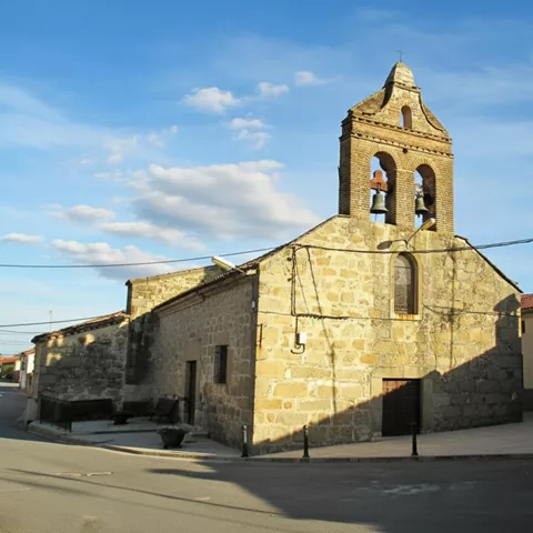 Vista frontal de una construcción religiosa antigua hecha de bloques de piedra, con una espadaña de ladrillo de dos niveles y una pequeña plaza empedrada en la base.