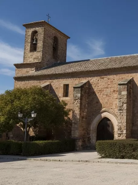 Fachada principal de la iglesia de Villapalacios, edificio histórico de piedra con contrafuertes y campanario bajo cielo azul.