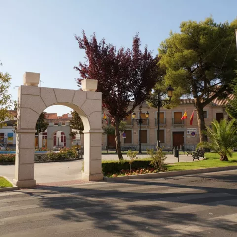 Plaza pública con un arco de piedra moderno, árboles frondosos, bancos de parque y edificios de ladrillo de fondo en un día soleado.