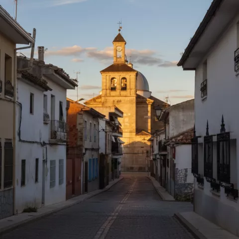 Una calle empedrada que se estrecha entre casas encaladas hacia una torre de iglesia bañada por el sol en primer plano.