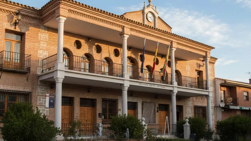 Una toma frontal captura un gran edificio municipal de ladrillo con dos balcones, columnas blancas y banderas, bajo un cielo matutino con nubes ligeras.