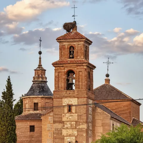 Primer plano de un campanario cuadrado de ladrillo con campanas visibles, coronado por un nido de cigüeña y una cruz.