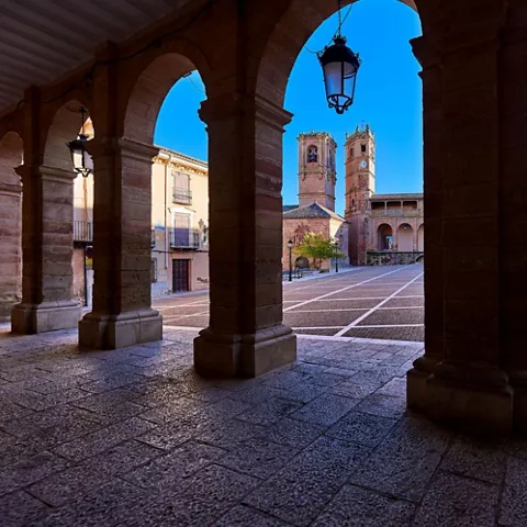 Plaza porticada con arcos de piedra y torres al fondo.