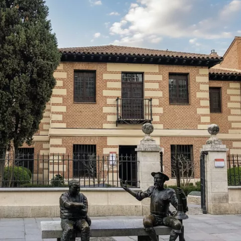 Fachada histórica con banco y esculturas de bronce frente a un edificio de ladrillo.