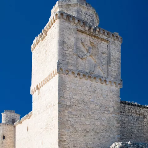 Torre de piedra con relieve tallado y muralla bajo cielo azul intenso.