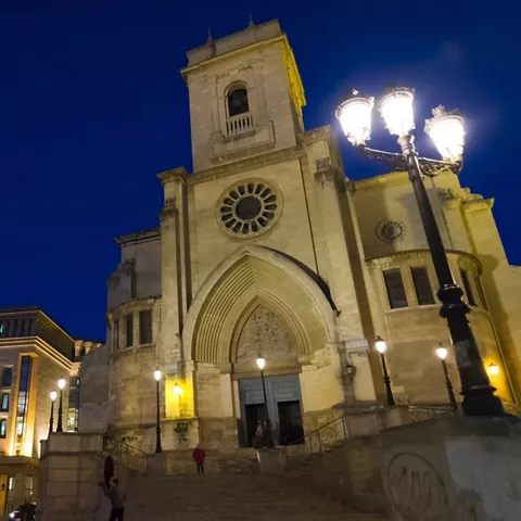 Fachada nocturna de iglesia iluminada con farolas y edificios urbanos alrededor.