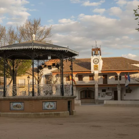 Quiosco de música en el centro de una plaza con edificio porticado y reloj.