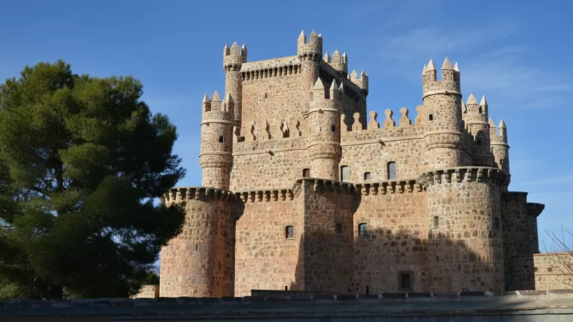 Un primer plano de un castillo medieval de piedra con múltiples torres circulares y almenas bajo un cielo azul intenso.