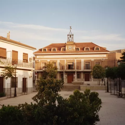 Una plaza de pueblo con edificios blancos a un lado y un edificio gubernamental con tejado de tejas rojas y un reloj en el centro, con árboles y farolas.