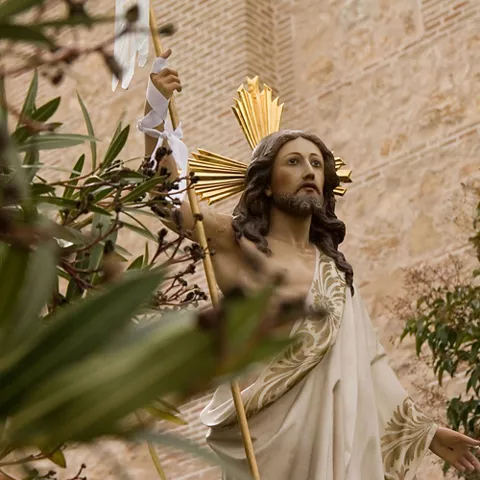 Escultura religiosa con halo dorado y estandarte, rodeada de vegetación.
