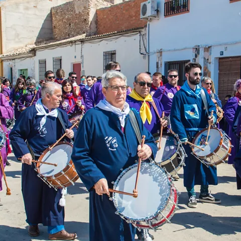 Grupo numeroso de personas tocando tambores durante una procesión diurna.