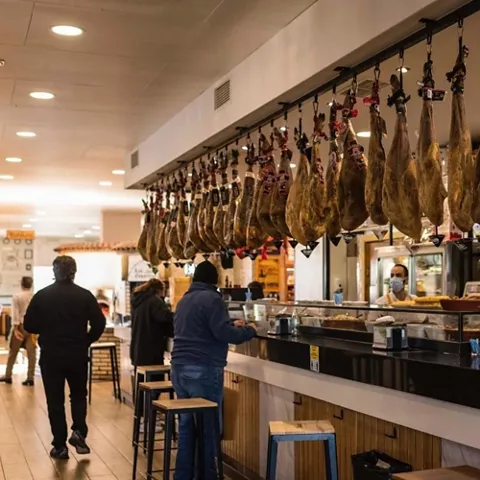 Bar con jamones colgados sobre la barra y clientes consumiendo.