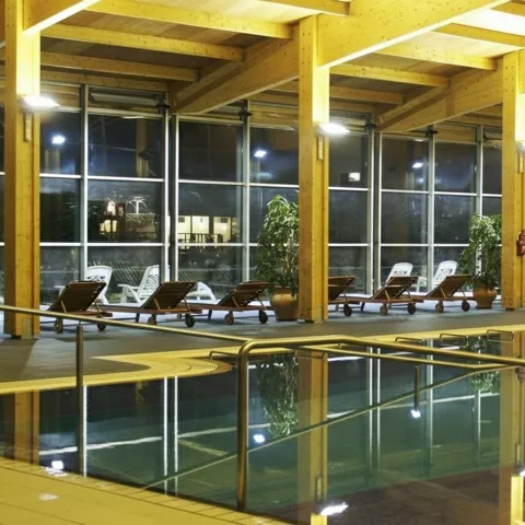 Piscina interior con estructura de madera, ventanales y fila de tumbonas junto al agua.