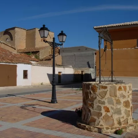 Plaza con pozo de piedra en primer plano y farolas, con edificios al fondo.