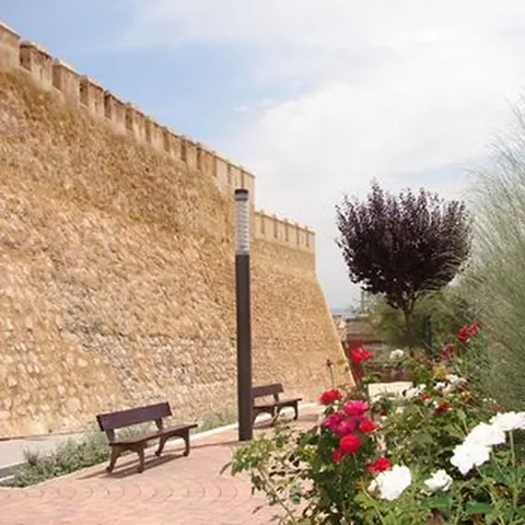Paseo ajardinado con bancos junto a una muralla histórica.
