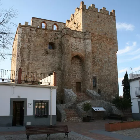 Edificio defensivo de piedra en plaza, con bancos y casas blancas alrededor.