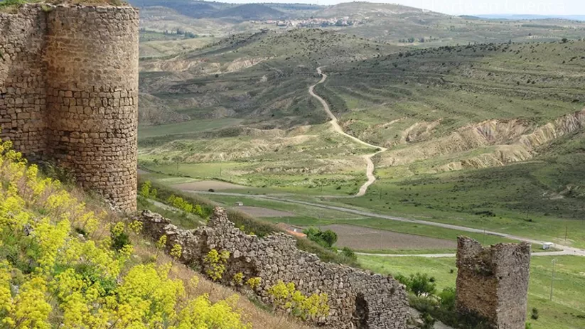 Restos de muralla y torre de piedra en una ladera, con caminos y campos extendiéndose al fondo.