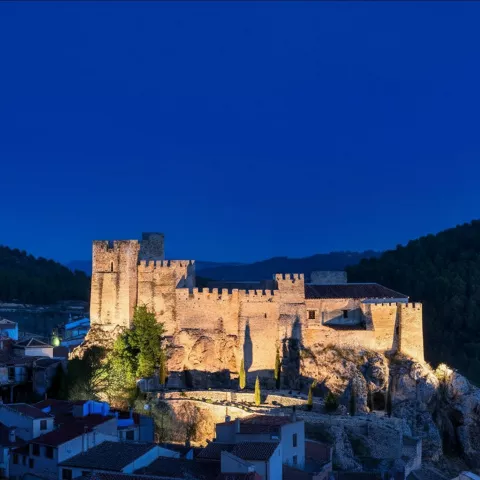 Castillo iluminado al anochecer sobre colina rodeada de casas.