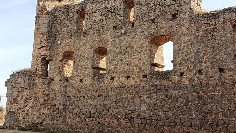 Ruinas de fortaleza de piedra con muros altos y ventanas en arco sobre terreno seco.