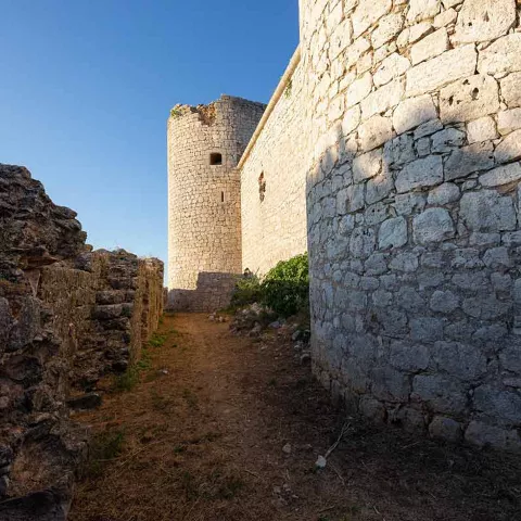 Camino de tierra entre murallas de piedra con torre defensiva al fondo bajo cielo despejado.
