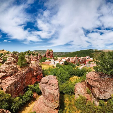 Formaciones rocosas rojizas con un pueblo al fondo y vegetación abundante.
