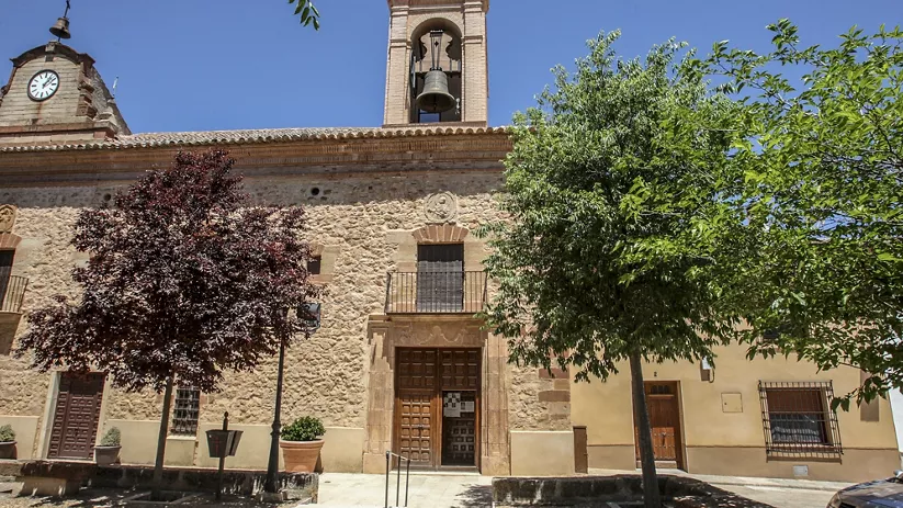 Fachada de iglesia de piedra con campanario y árboles en la plaza.