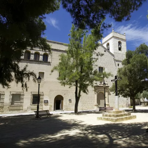 Fachada de iglesia de piedra con torre campanario y plaza arbolada.