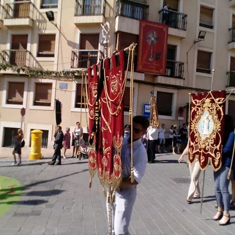 Persona porta un estandarte rojo bordado en una plaza, con público al fondo.