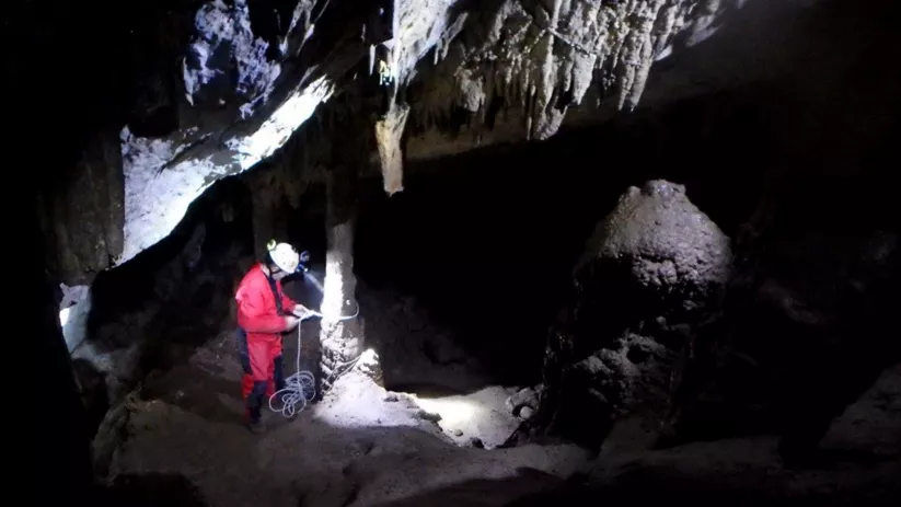 Espeleólogo con casco y cuerda bajo estalactitas en una sala oscura de cueva.