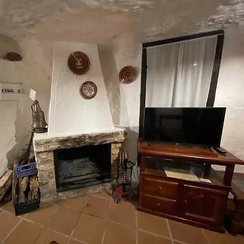 Salón de casa cueva con chimenea de piedra, leña apilada y mueble con televisor.