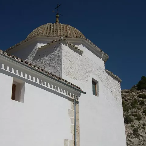 Detalle de cúpula de ermita blanca con tejas curvas y veleta sobre cielo azul.